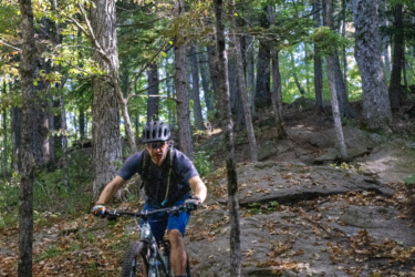 A mountain biker navigating a rocky trail surrounded by trees with vibrant autumn foliage. The biker is wearing a helmet and preparing to tackle the uneven terrain as leaves cover the ground. Mt. Peg mountain bike trail.