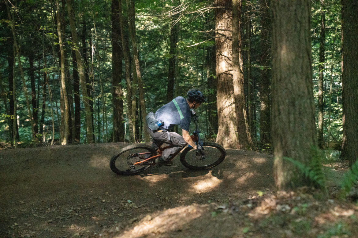 A mountain biker leaning into a turn on a dirt path surrounded by tall trees in a wooded area. Sunlight filters through the leaves, creating dappled shadows on the ground. The biker is wearing a helmet and casual cycling attire.