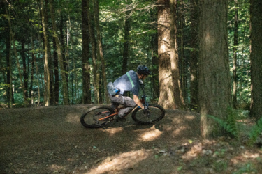 A mountain biker navigating a dirt path in a wooded area, leaning into a turn as sunlight filters through the trees. The cyclist is wearing a helmet and riding a bike with an orange frame, surrounded by lush green vegetation and tall trees. Mt. Peg mountain bike trail.
