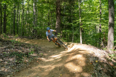 A mountain biker leans into a turn on a dirt trail surrounded by lush green trees, showcasing a dynamic riding posture on a sunny day. Silver Moose mountain bike trail.