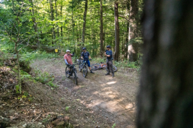 Three mountain bikers pause on a dirt trail surrounded by lush green trees. One rider is gesturing while the others listen, with their bikes parked nearby. Sunlight filters through the foliage, illuminating the scene in a vibrant outdoor setting. Mt. Peg mountain bike trail.