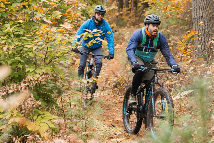 Two mountain bikers navigate a trail surrounded by autumn foliage. The riders are wearing helmets and outdoor gear, with one in a blue jacket and the other in a black and green outfit. The path is lined with vibrant orange and yellow leaves, creating a colorful fall scene.