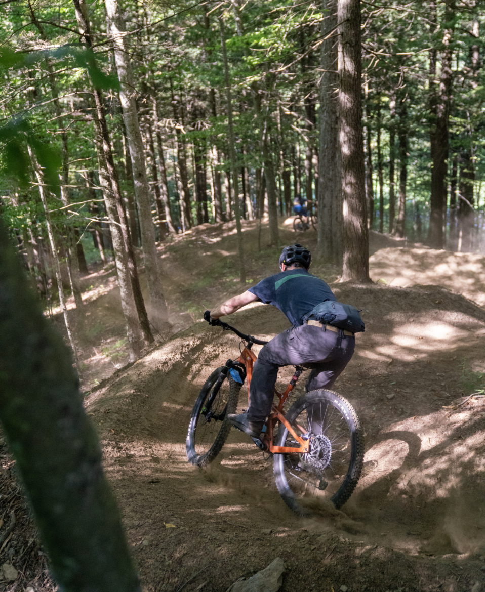 A mountain biker navigates a dirt trail in a forest, leaning into a turn as dust kicks up from the tires. Trees surround the path, creating a lush green backdrop, while another biker is visible in the distance.
