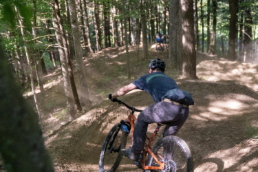 A person biking down a dirt trail in a forest, surrounded by tall trees. The cyclist is leaning into a turn on a mountain bike, with dust kicking up from the tires. Another biker can be seen in the background. The scene captures the essence of outdoor mountain biking in a natural setting. Mt. Peg mountain bike trail.