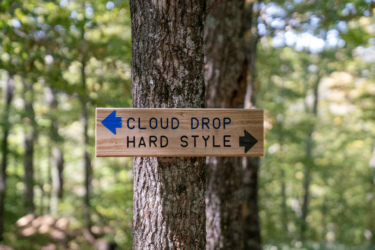 Wooden directional sign mounted on a tree in a forest, indicating two paths: "Cloud Drop" to the left and "Hard Style" to the right, with blue and black arrows pointing in the respective directions. Background features blurred greenery and trees. Mt. Peg mountain bike trail.