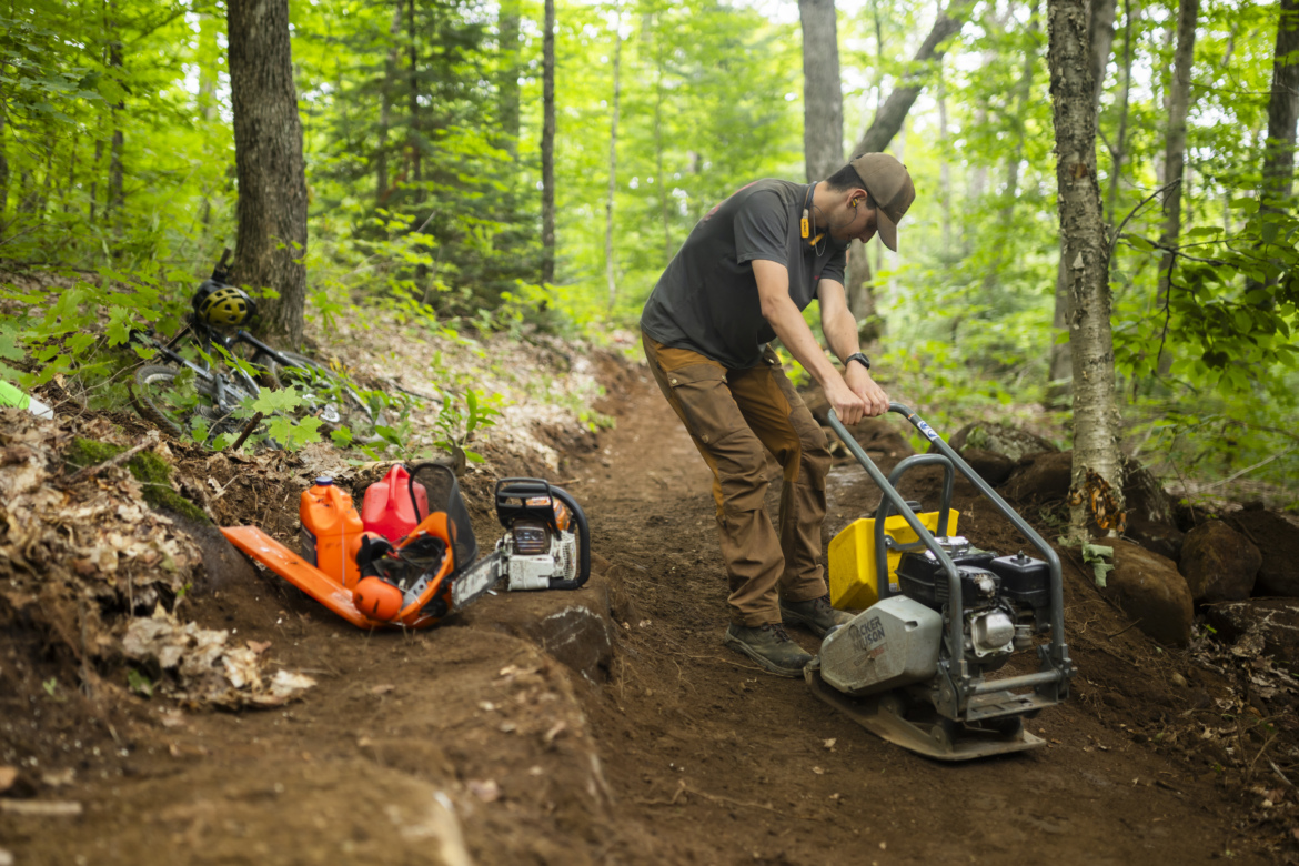 A person wearing a cap and work clothing operates a tamper machine on a dirt path in a forested area. Nearby, tools including a chainsaw and fuel canisters are placed on the ground. A mountain bike with a helmet is partially visible to the side, and the surrounding landscape is lush with green foliage and trees.