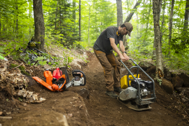 A person wearing a cap and work clothing operates a tamper machine on a dirt path in a forested area. Nearby, tools including a chainsaw and fuel canisters are placed on the ground. A mountain bike with a helmet is partially visible to the side, and the surrounding landscape is lush with green foliage and trees.