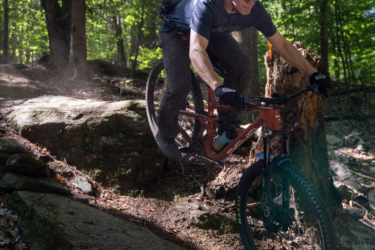 A mountain biker navigating a rocky trail in a wooded area, leaning forward on their bike as they approach a large boulder. Sunlight filters through the trees, casting dappled light on the ground. Dust kicks up from the tires as the rider skillfully maneuvers over the uneven terrain. Mt. Peg mountain bike trail.