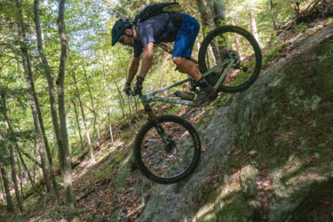 A mountain biker navigating a rocky downhill trail in a lush, green forest during daylight. The rider is wearing a helmet and sporty gear, leaning forward on the bike as they balance on a large rock surrounded by trees and foliage. Mt. Peg mountain bike trail.
