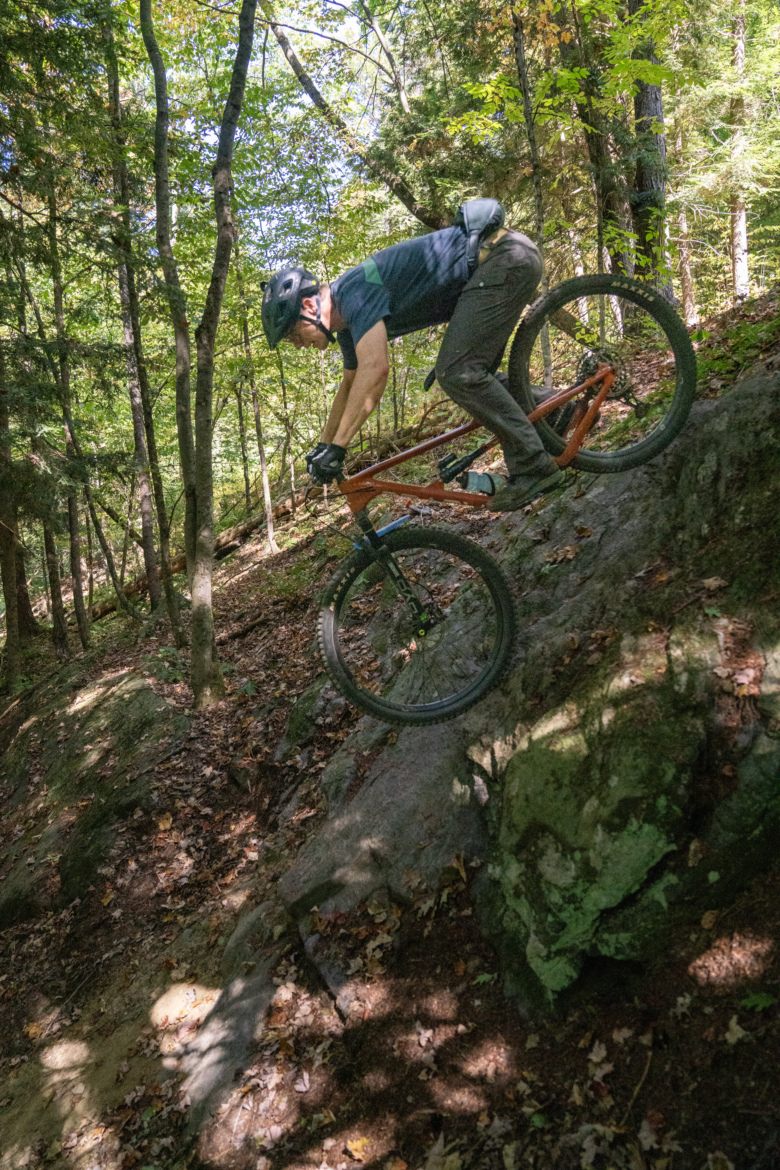 A mountain biker descending a rocky slope in a forested area, surrounded by trees and fallen leaves, wearing a helmet and outdoor gear.