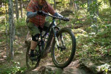 A mountain biker navigating rocky terrain in a forested area, wearing a plaid shirt and shorts, with a focused expression. Dust is kicked up from the bicycle wheels as the rider maneuvers over the rocks surrounded by greenery and trees. Mt. Peg mountain bike trail.
