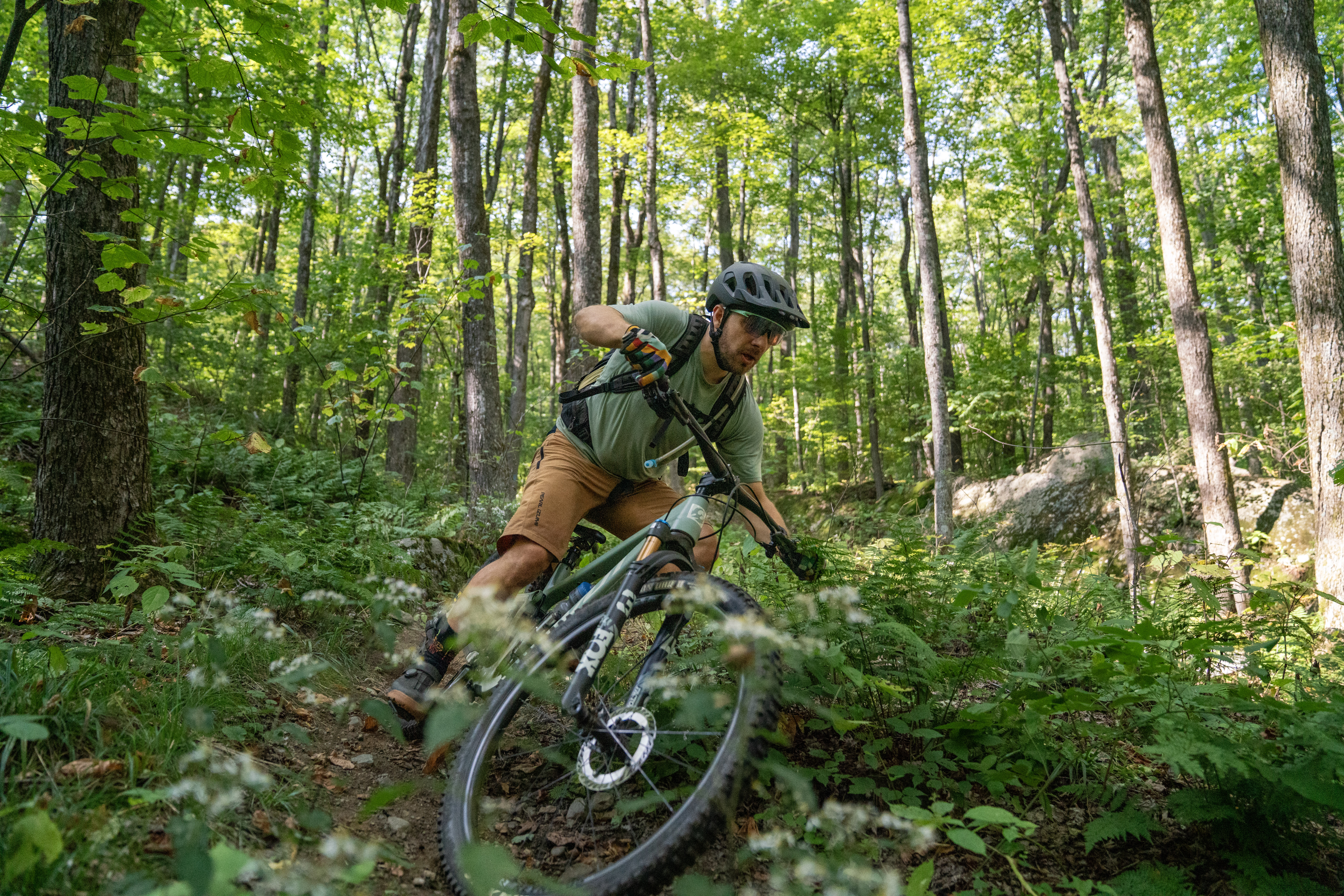 A mountain biker navigating a narrow, winding trail through a lush forest. The rider, wearing a helmet and gloves, leans into a turn as the bike