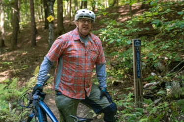 A person wearing a helmet and glasses stands next to a mountain bike on a wooded trail, with a signpost labeled "KENT'S CONNECTOR" visible in the background. The individual is dressed in a plaid shirt and shorts, surrounded by green foliage and trees. Mt. Peg mountain bike trail.