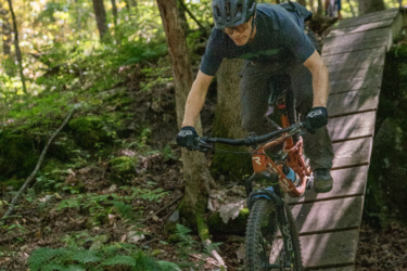 A mountain biker riding a wooden bridge over a forested area, surrounded by trees and ferns, with a second biker in the background. The rider is focused and wearing a helmet, navigating the narrow path. The scene captures the essence of outdoor adventure and mountain biking. Mt. Peg mountain bike trail.