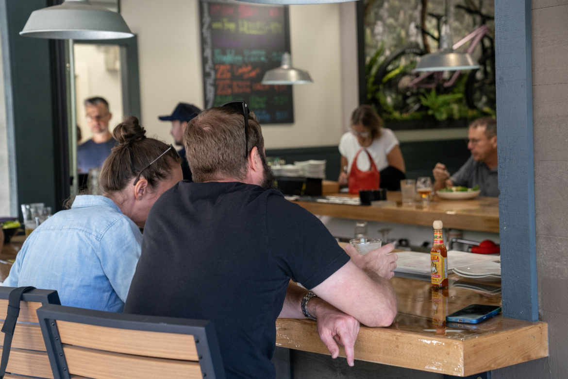 A couple sits at a bar in a restaurant, with their backs to the camera. The woman wears a light blue denim jacket and has her hair in a bun, while the man is wearing a black shirt and has a beard. The scene is lively, with a few other diners visible in the background, and a variety of food and drinks on the counter. A bottle of hot sauce and a phone are on the bar in front of them.