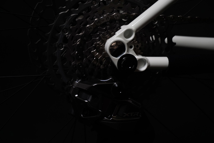 Close-up view of a bicycle's rear drivetrain, featuring a white bicycle frame section, a black derailleur, and a metallic cassette, all set against a dark background.