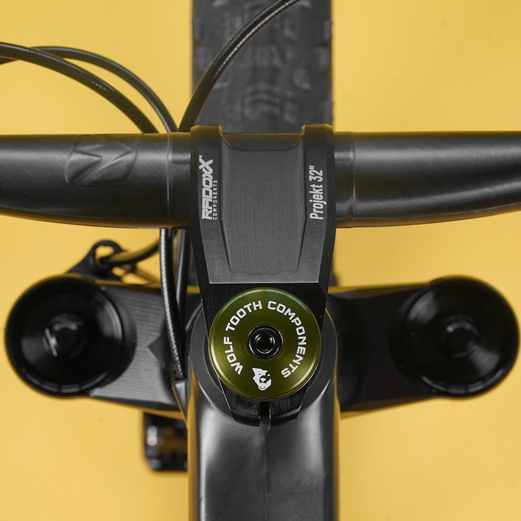 Close-up view of a bicycle handlebar and stem featuring a black and green design. The stem includes branding for "Wolf Tooth Components" and "Projekt 32," with cables neatly routed alongside. The background is a vibrant yellow, enhancing the focus on the bike components.