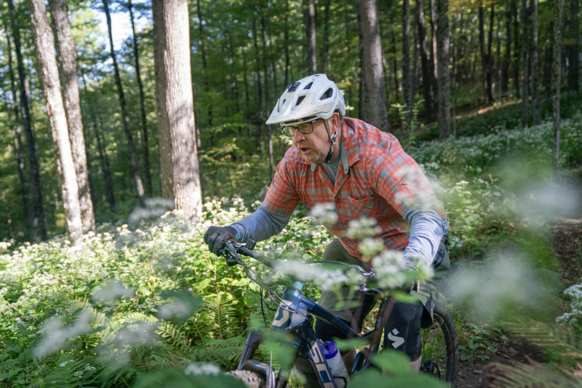 An older man wearing glasses and a helmet rides a mountain bike through a lush green forest trail, surrounded by greenery and sunlight filtering through the trees. He is dressed in a plaid shirt and appears focused on the ride.