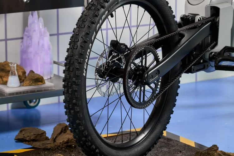 A close-up of a mountain bike's rear tire positioned on a dirt patch with rocks, set against a colorful laboratory backdrop featuring glassware and purple elements. The tire has a rugged tread pattern and is mounted on a modern bike frame. The scene includes a yellow and black striped border around the dirt area, enhancing the visual contrast.