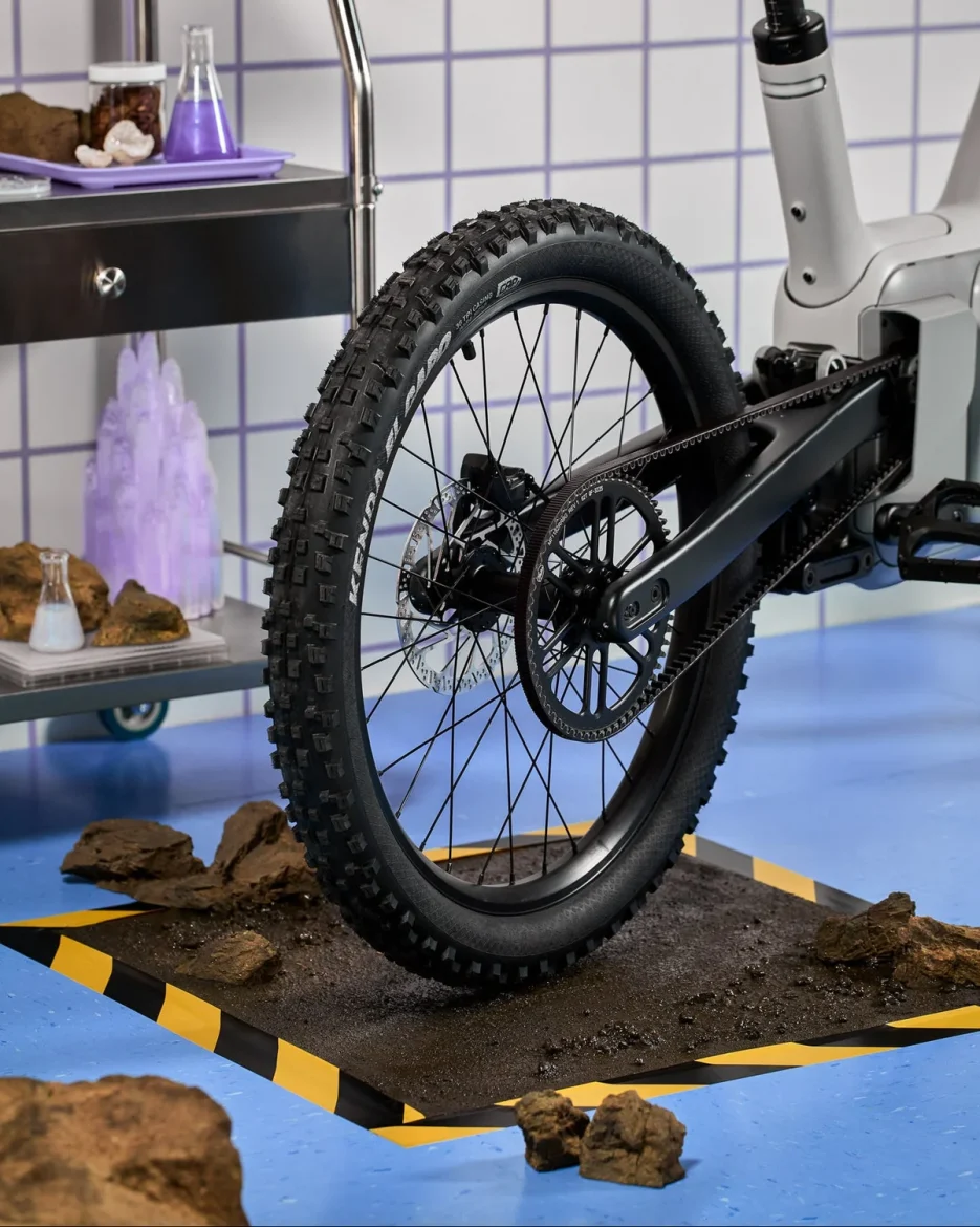 A close-up of a mountain bike's rear tire positioned on a dirt patch with rocks, set against a colorful laboratory backdrop featuring glassware and purple elements. The tire has a rugged tread pattern and is mounted on a modern bike frame. The scene includes a yellow and black striped border around the dirt area, enhancing the visual contrast.