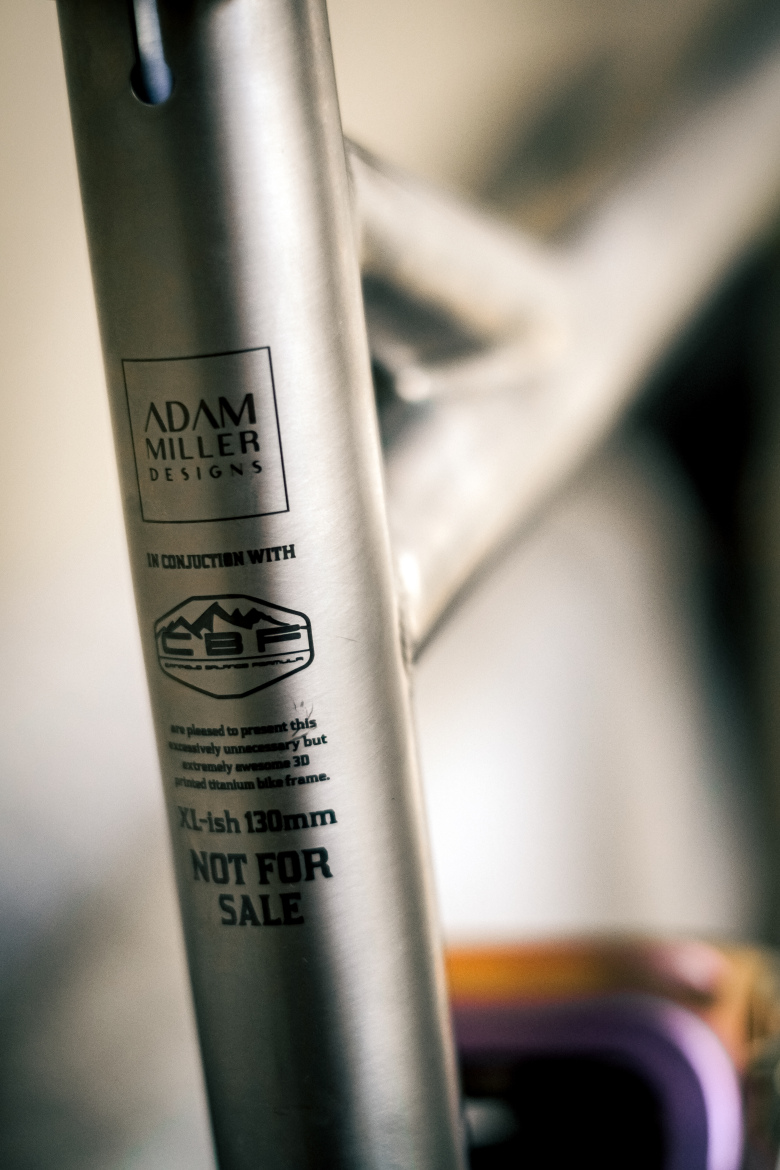 Close-up image of a titanium bike frame featuring branding from "Adam Miller Designs" and "CBF." The frame is labeled with details stating "XL-ish 130mm" and "NOT FOR SALE." The text emphasizes a unique design and highlights the frame's 3D printed technology.