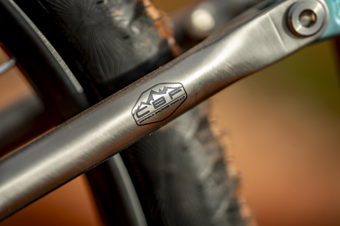 Close-up image of a bicycle's crank arm featuring the "CBF" logo, with the background blurred and showing part of a tire. The metal surface of the crank arm is polished, and there is some dirt visible, suggesting use in an outdoor environment.