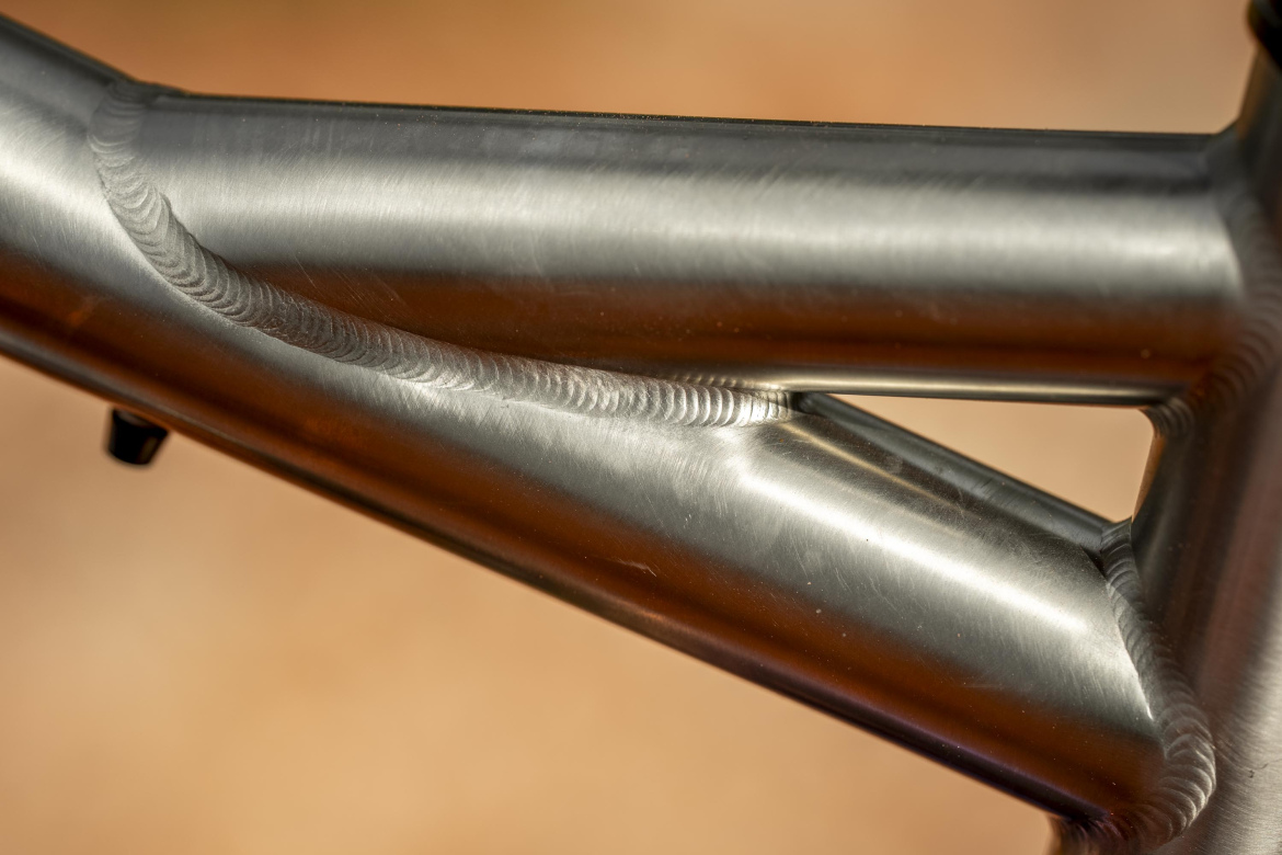 Close-up image of a metal bicycle frame highlighting a smooth, polished weld seam. The focus is on the intricate details of the welding and the texture of the metal.