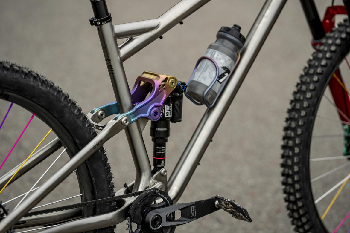 A close-up view of the rear suspension and drivetrain of a mountain bike. The bike features a metallic frame, a colorful shock linkage component, and a water bottle secured in a holder. The background shows a blurred surface.