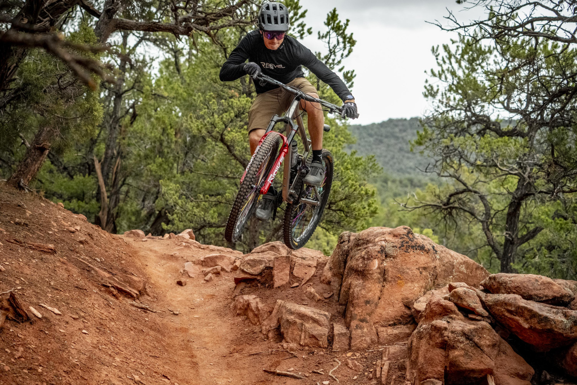A mountain biker performing a jump on a rocky trail surrounded by trees. The rider wears a helmet and protective gear, showcasing an action-packed moment in nature.