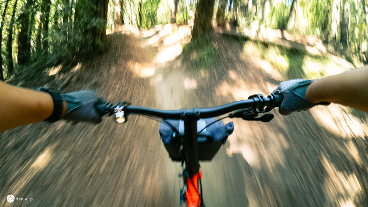 A cyclist's perspective, with hands gripping the bike handlebars on a dirt path surrounded by greenery, capturing a sense of speed and movement through a forested trail.