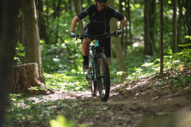 An individual riding a mountain bike on a winding trail in a lush green forest, surrounded by trees and foliage. The rider is focused, wearing a helmet and gloves, as sunlight filters through the leaves, illuminating the path ahead. Durham Forest mountain bike trail.