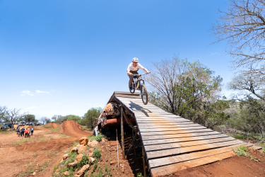 A mountain biker performing a jump off a wooden ramp against a bright blue sky. In the background, there are spectators watching and dirt biking trails visible. The surroundings include trees and rocky terrain. Reveille Peak Ranch mountain bike trail.