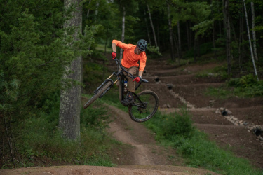 A mountain biker wearing an orange shirt and gloves performs a jump on a dirt trail surrounded by trees. The rider is mid-air with the bike angled to one side, showcasing action and excitement in a natural outdoor setting. Mt. Telemark mountain bike trail.