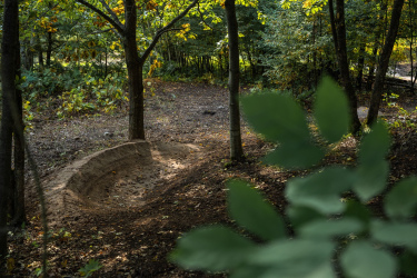 A dirt mountain bike track winding through a forested area, lined by trees with green and yellow leaves. The track features a curved jump section, and the ground is covered with scattered leaves and soft earth. Sunlight filters through the trees, creating a dappled light effect on the path. Mt. Telemark mountain bike trail.