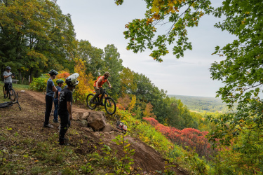 A group of mountain bikers observe as one rider jumps off a rock formation on a scenic trail surrounded by vibrant autumn foliage. In the background, a panoramic view of hills and trees stretches out under a cloudy sky. Mt. Telemark mountain bike trail.