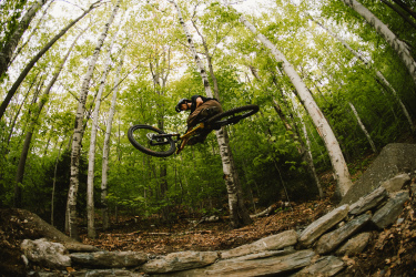 A mountain biker performs a jump over a rocky ramp in a lush green forest, surrounded by tall trees with vibrant leaves. The rider is mid-air, showcasing an impressive trick as they navigate the trail. Purple Valley Trails mountain bike trail.
