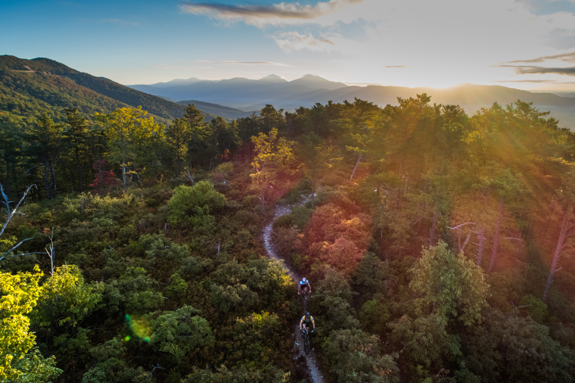 A scenic view of a winding mountain trail surrounded by lush greenery, with two mountain bikers riding through the foliage. The background showcases distant mountains under a clear sky, illuminated by the warm glow of a sunset, highlighting the vibrant colors of autumn foliage in the foreground.