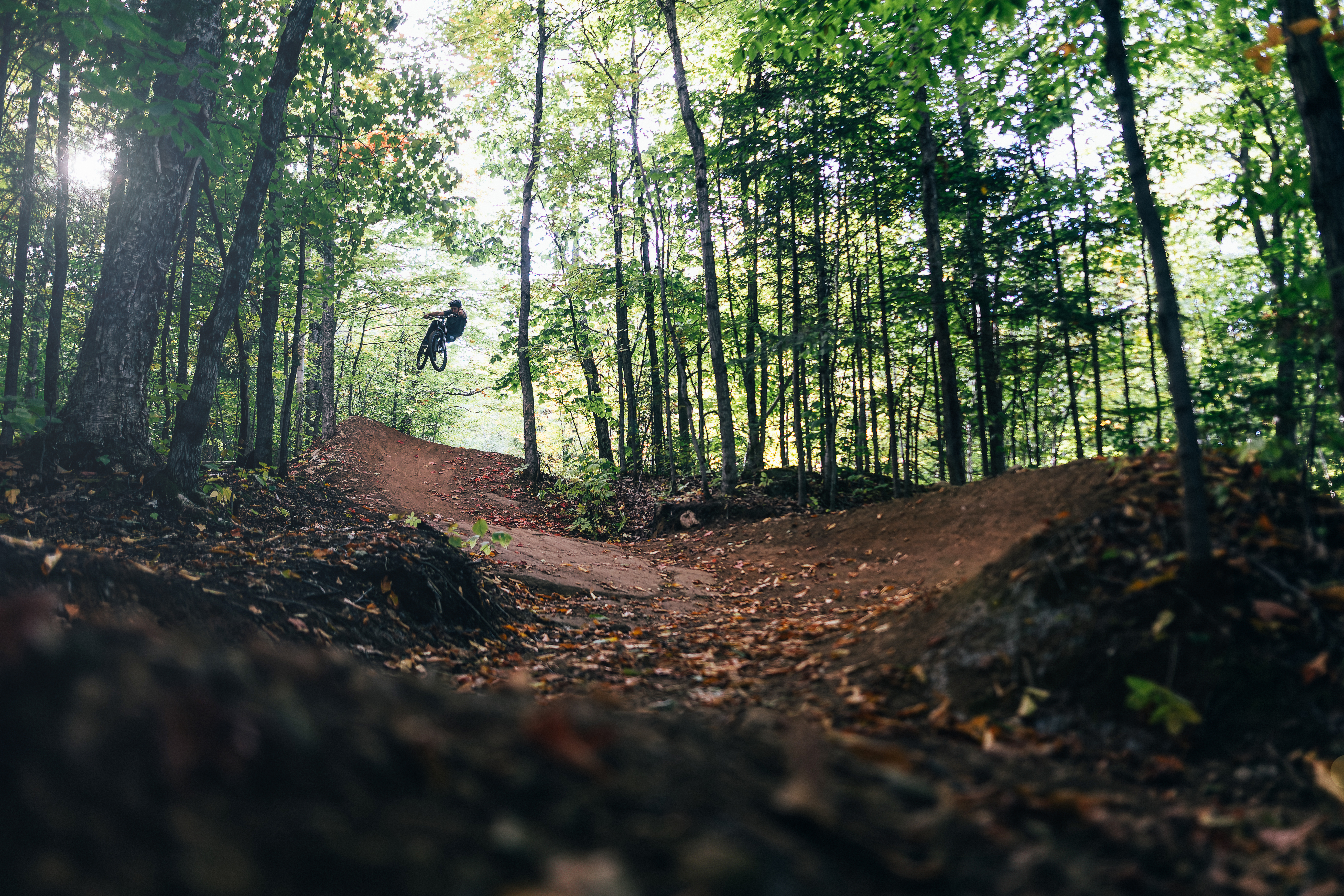 A mountain biker performing a jump in a forested area, surrounded by trees with green leaves. The dirt trail features an elevated jump ramp in the foreground. Mont SM mountain bike trail.