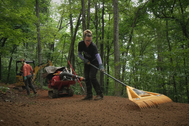 A woman using a rake to smooth freshly tilled soil in a wooded area, with trees in the background. Another person is seen operating a small excavator nearby, while a red wheelbarrow is positioned in the foreground. The scene captures an active outdoor work environment focused on land preparation. Aetna Mountain mountain bike trail.