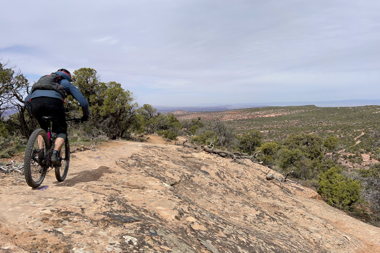 A mountain biker rides along a rocky trail with a vast, scenic landscape in the background, showcasing green shrubs and distant hills under a cloudy sky.
