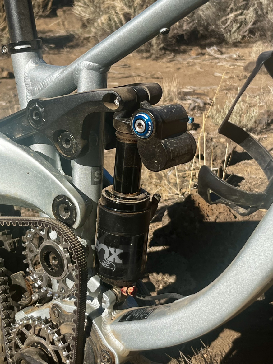 Close-up image of a mountain bike suspension system, featuring a Fox Float shock and aluminum frame components. The photo showcases the intricate details of the bike's design, including the gear mechanism and linkage system, set against a natural outdoor background.
