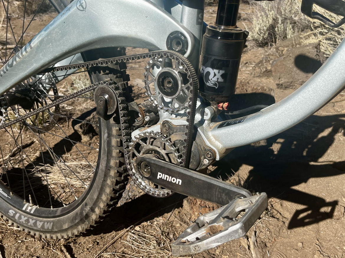 Close-up view of a mountain bike's drivetrain, showcasing the crankset, chainring, and pedal, with a visible pinion gear system. The background features rocky terrain and sparse vegetation, indicating an outdoor setting.