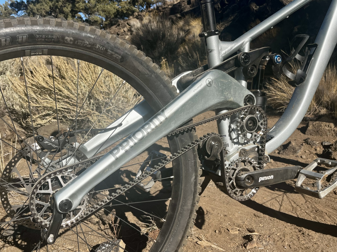 Close-up view of the drivetrain and rear wheel of a mountain bike, showcasing the gear components, chain, and brake disc against a background of rocky terrain and sparse vegetation. The bike features a Pinion gear system and is positioned in natural outdoor surroundings.
