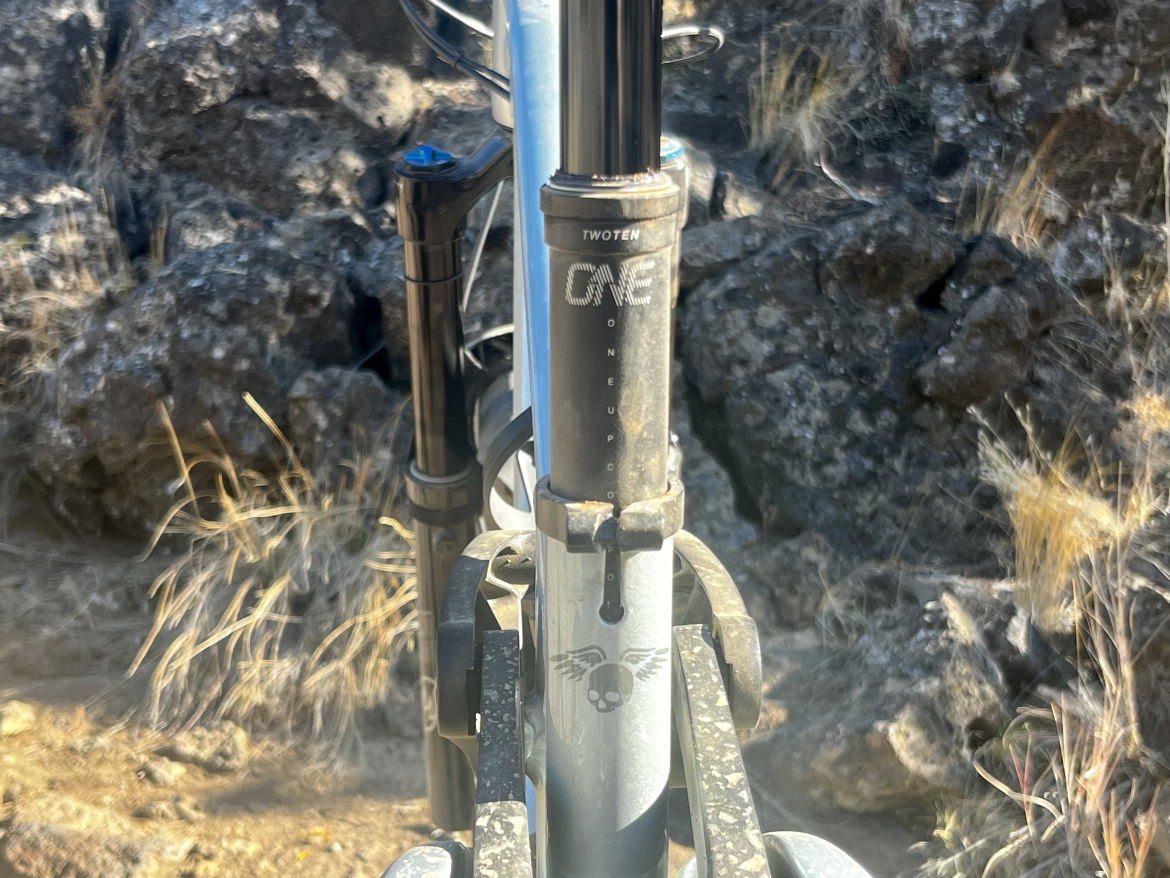 Close-up view of a mountain bike fork with visible branding and details, set against a rocky landscape and dry grass. The focus is on the fork's components, showcasing its rugged design and wear.