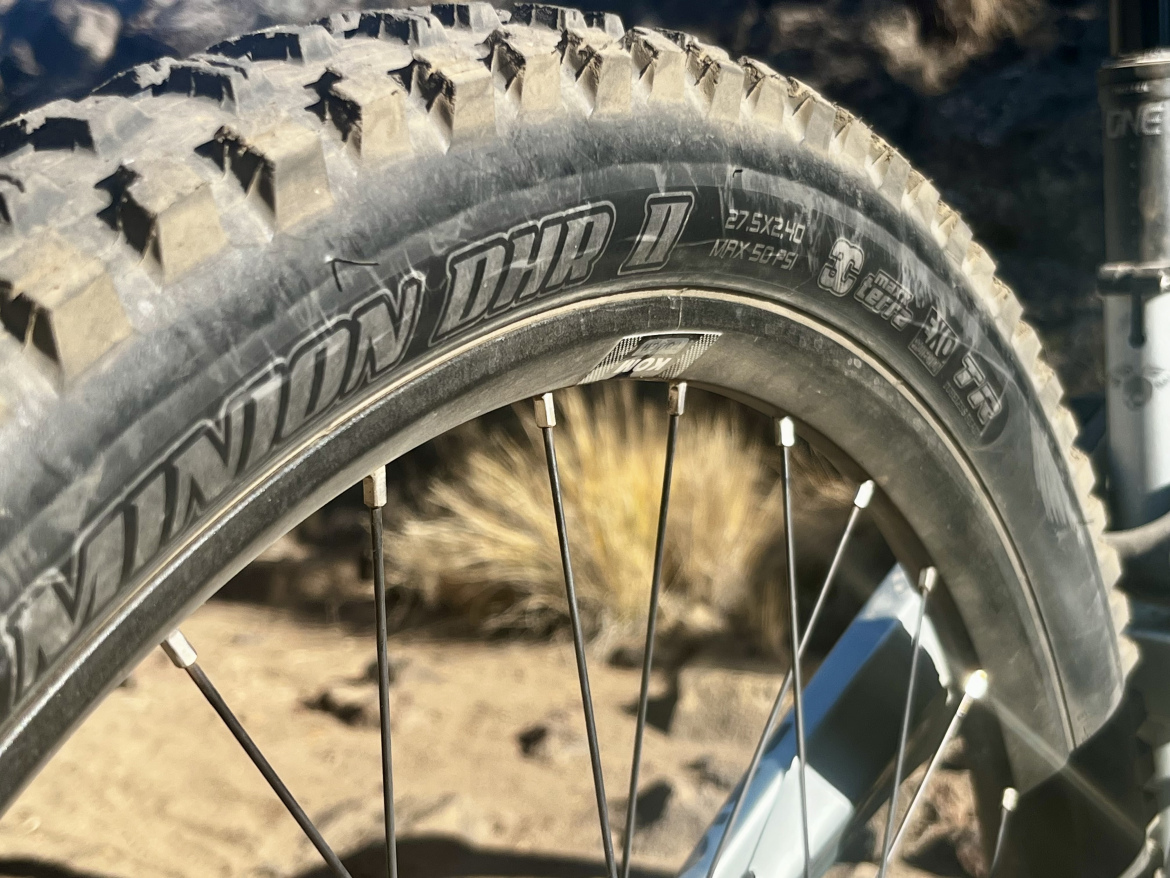  close-up view of a mountain bike tire with prominent tread patterns, showcasing the branding "Minion DHR II". The tire size is visible as 27.5 x 2.40, set against a natural background of rocky terrain and dry vegetation.