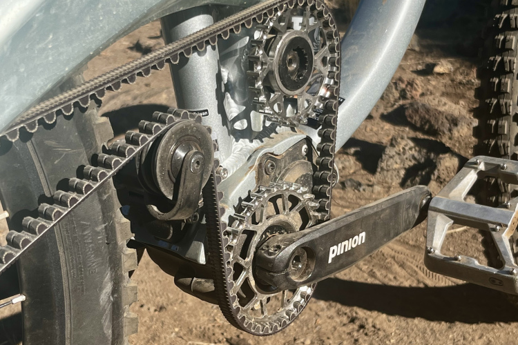 Close-up image of a bicycle's drivetrain, featuring a pinion gear system, pulleys, and a belt drive, set against a dirt background. The components show intricate details of the gears and chain, illustrating the mechanics of the bike's pedaling system.