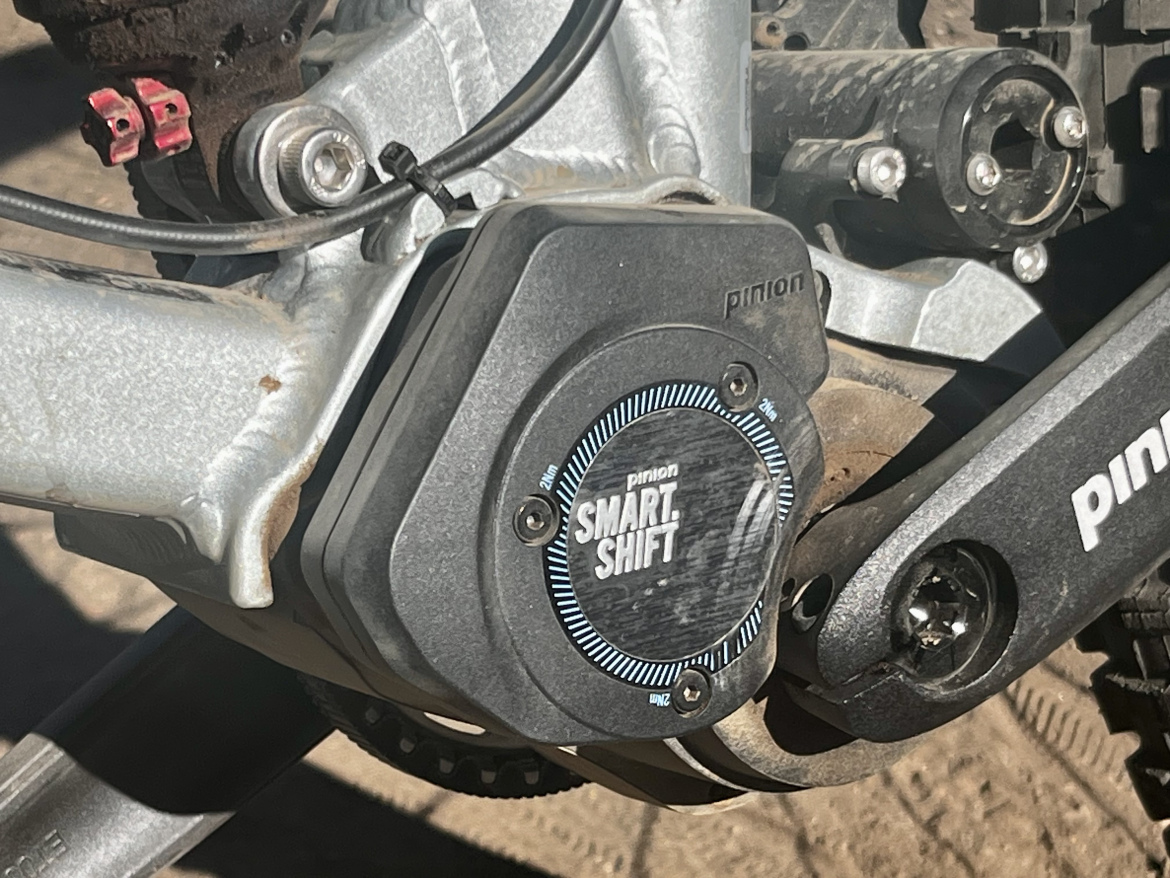 Close-up image of a Pinion Smart Shift gearbox mounted on a bicycle frame, showcasing the mechanical details, including wiring and connectors. The background features a sandy surface, indicating an outdoor setting.