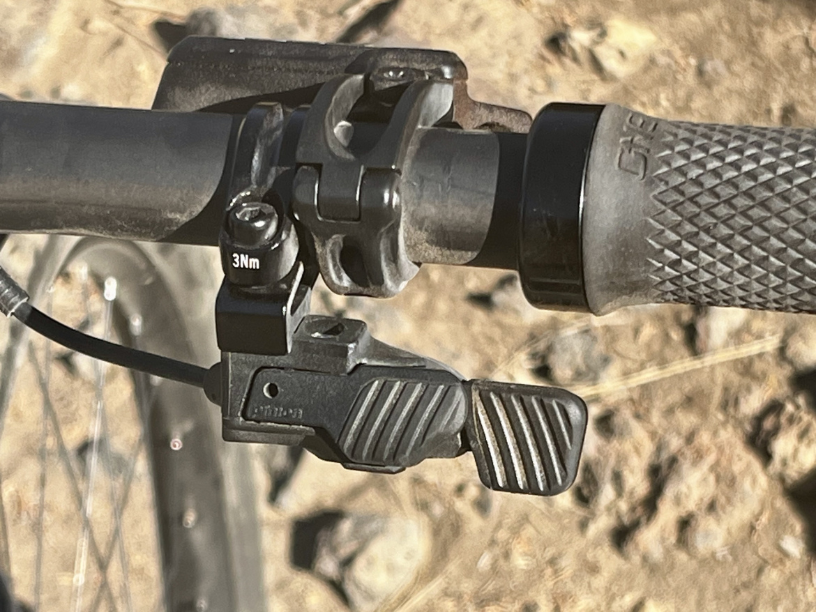Close-up view of a bicycle handlebar with a gear shift lever and brake lever attachments. The handlebar features textured grips and a visible cable connected to the shift lever, which has a torque specification indicator of 3Nm. The background shows a dirt surface, suggesting an outdoor setting.