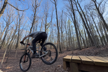 A mountain biker performing a jump off a wooden ramp in a forested area during the daytime. The scene features leafless trees and a clear blue sky in the background. Locust Shade Park mountain bike trail.