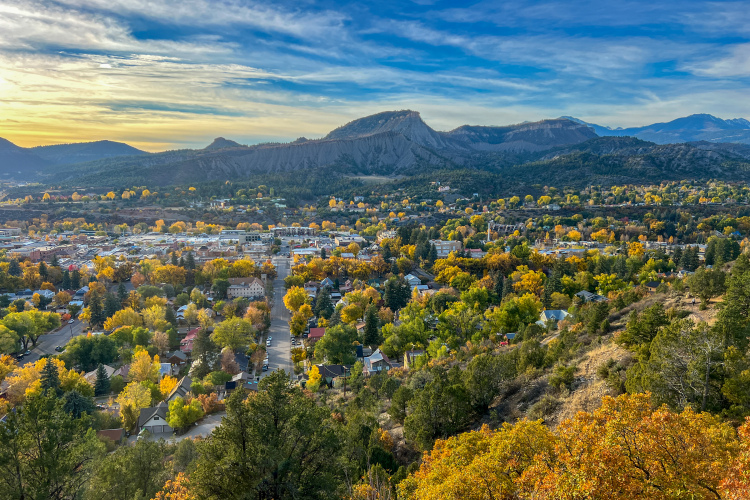 A panoramic view of a small town surrounded by colorful autumn foliage and mountains under a blue sky. The town features a mix of residential and commercial buildings, with trees displaying vibrant yellow, orange, and green leaves.